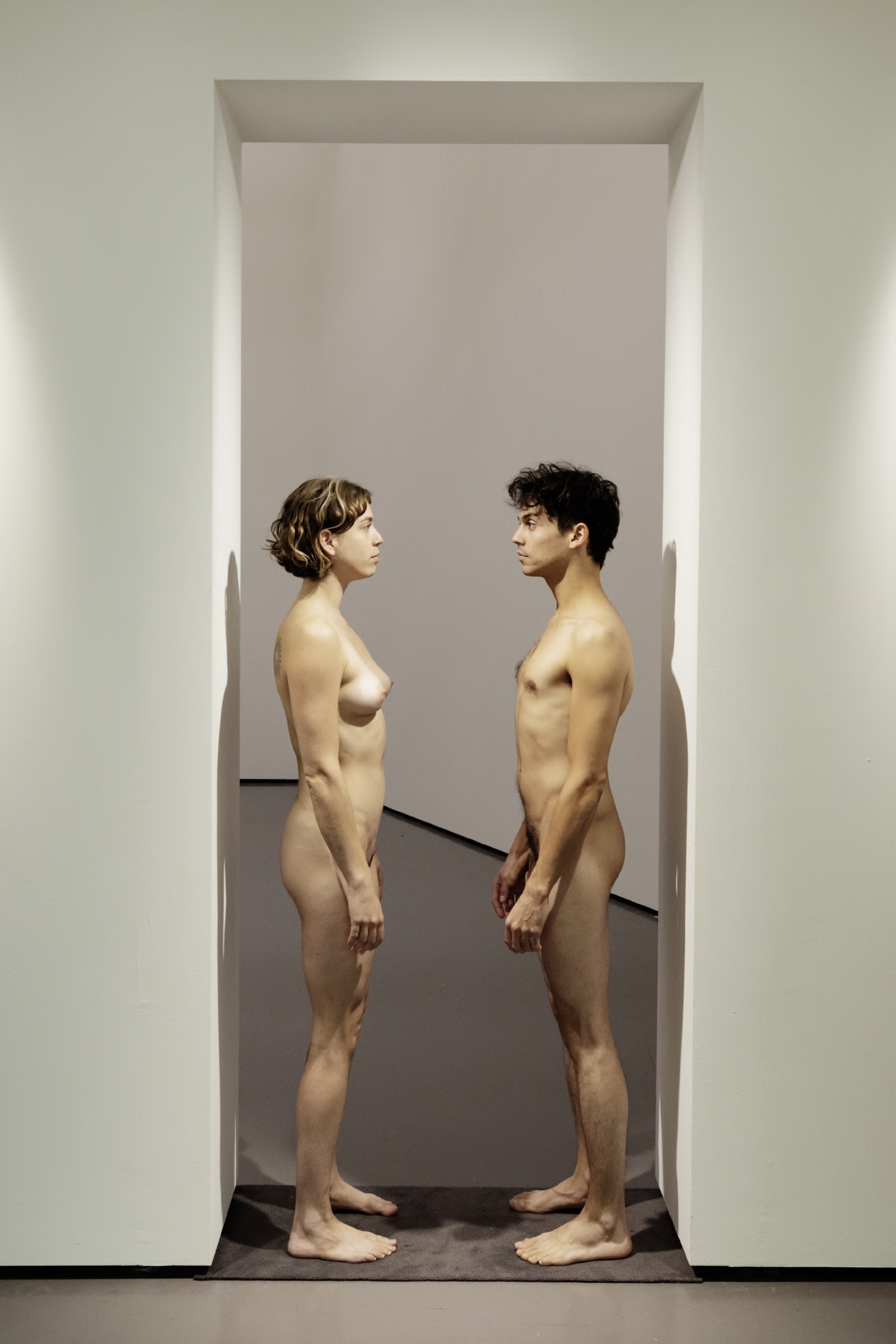 Two naked performers facing each other in a narrow doorway