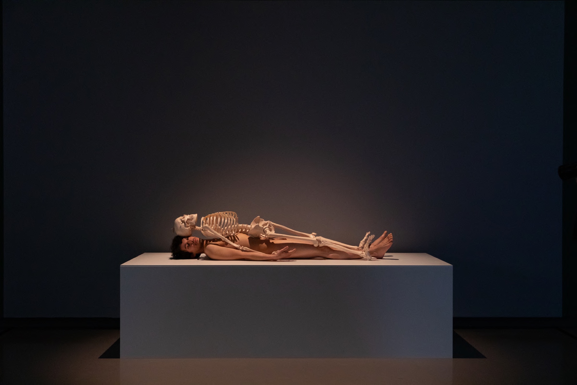 Performer lying under a skeleton on a white plinth