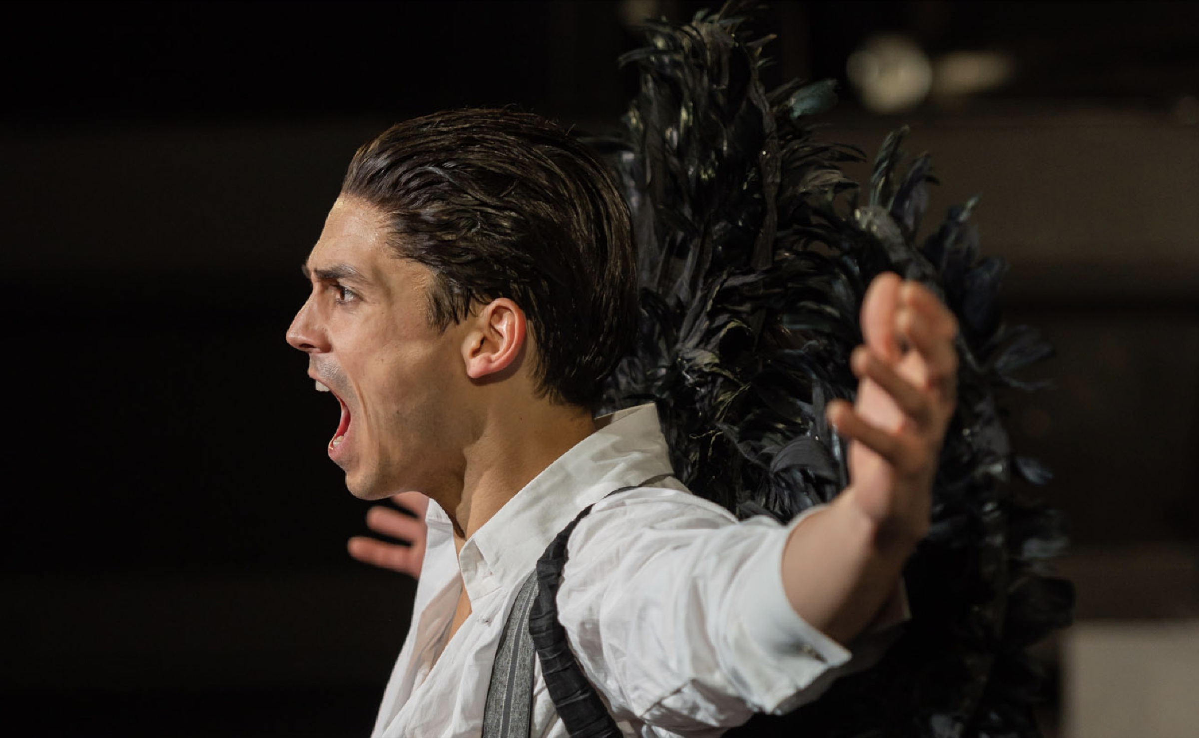 Actor in a white shirt shouting, black feathers behind him