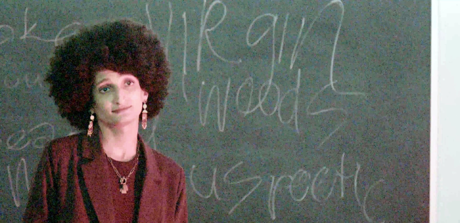 A performer with an afro and long earrings in front of a chalkboard