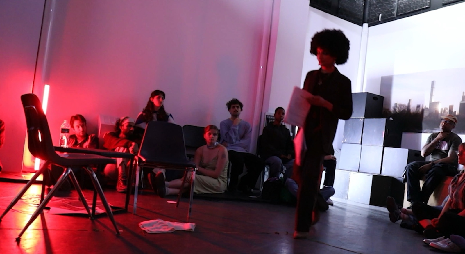The studio set: a red neon tube and a chair, stepped black cubes before a skyline print, the audience on the floor, a performer with an afro crossing the space