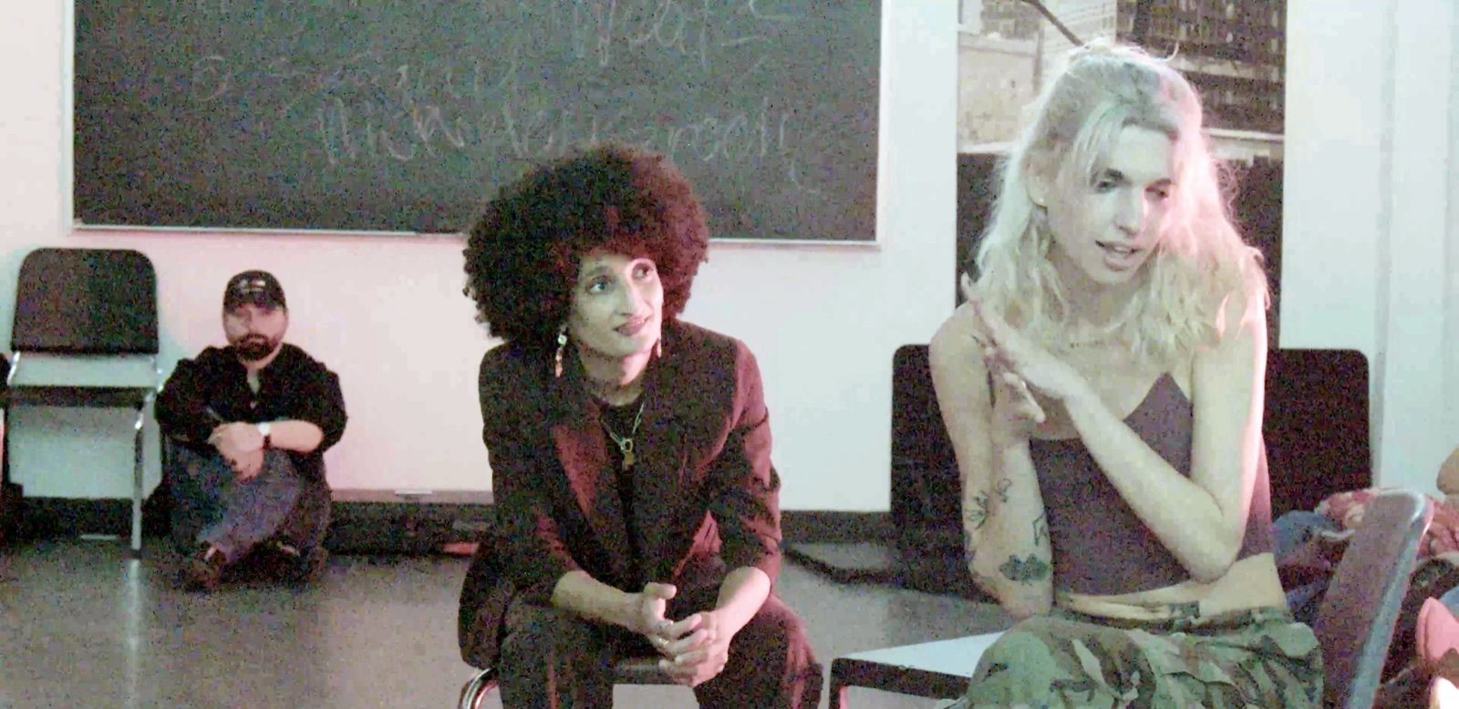 Two performers seated side by side, one with an afro and a dark red suit, one in a crop top and camouflage trousers